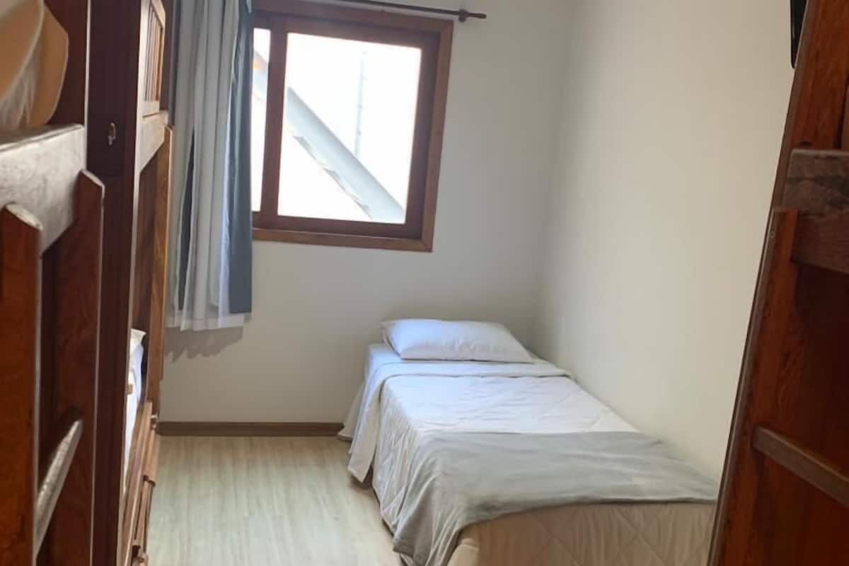 Une petite chambre avec un lit simple, un plancher en bois, une fenêtre avec des rideaux et un climatiseur fixé au plafond.