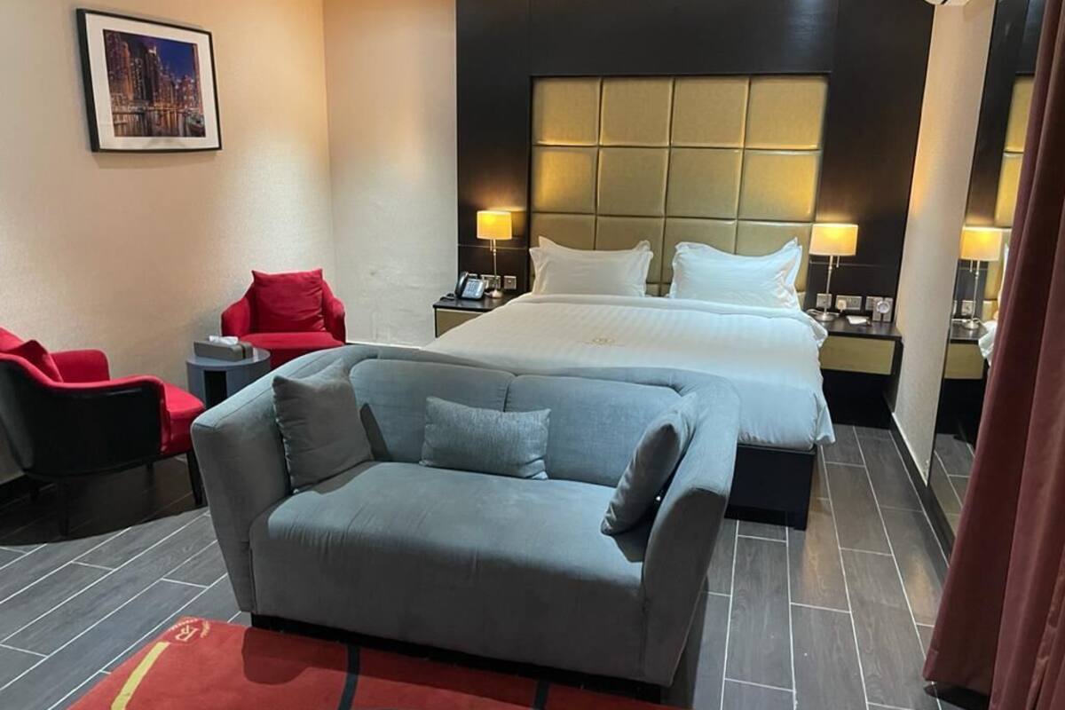 A hotel room with a large bed, a grey sofa, a red armchair, and a red rug.
