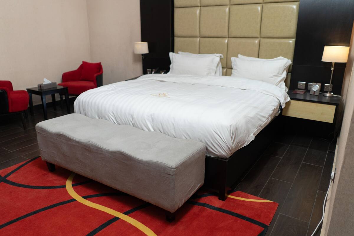 A hotel room with a large bed, a red chair, a small table, and a nightstand with a lamp.