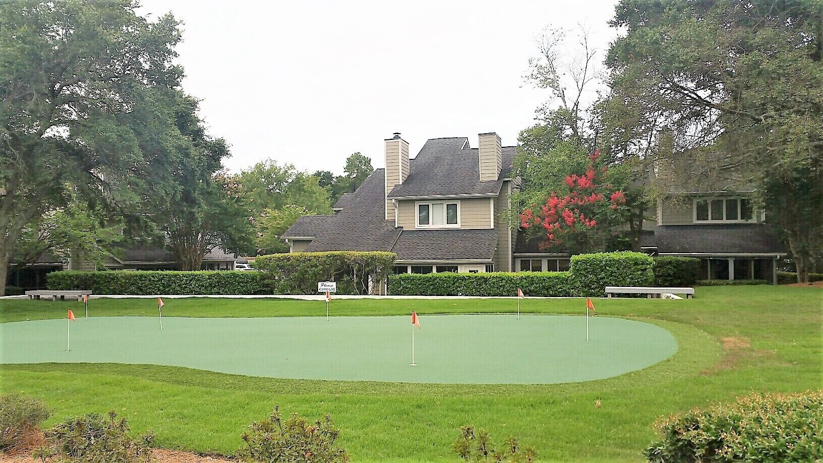 Campo de golfe com putting green, rodeado por relvados bem cuidados e sebes, com casas ao fundo.