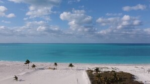 Ein Strand mit weißem Sand, türkisblauem Wasser und Palmen unter einem teilweise bewölkten Himmel.