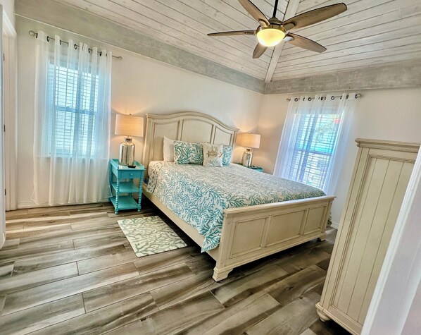 Une chambre avec un plancher en bois, un lit recouvert d’une literie à motifs, deux tables de chevet avec des lampes, un ventilateur de plafond et une fenêtre ornée de rideaux légers.
