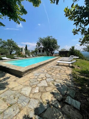 Ein Poolbereich mit Steinpflasterung, Liegestühlen und einem Haus im Hintergrund.