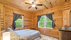 Habitación de cabaña de madera con cama, dos ventanas con cortinas, ventilador de techo y mesita de noche con lámpara.