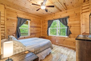 A wooden cabin room with a bed, two windows with curtains, a ceiling fan, and a nightstand with a lamp. - Double Down Lodge | 5BR Group Getaway, Hot Tub & Game Room Near Old Man's Cave (Logan)