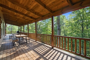 Una terraza de madera techada con mesas de picnic y vistas a un bosque.