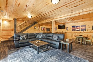 A log cabin interior with a black leather sofa, wooden coffee table, and a staircase. - Double Down Lodge | 5BR Group Getaway, Hot Tub & Game Room Near Old Man's Cave (Logan)