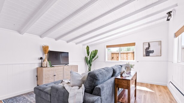 Ein modernes Wohnzimmer mit einem grauen Ecksofa, einem Holzkabinett, einem Flachbildfernseher und einem großen Fenster mit Blick ins Freie.