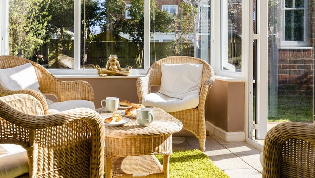 Een zonnige kamer met rieten meubels, een tafel met gebak en uitzicht op een tuin en huizen door grote ramen.