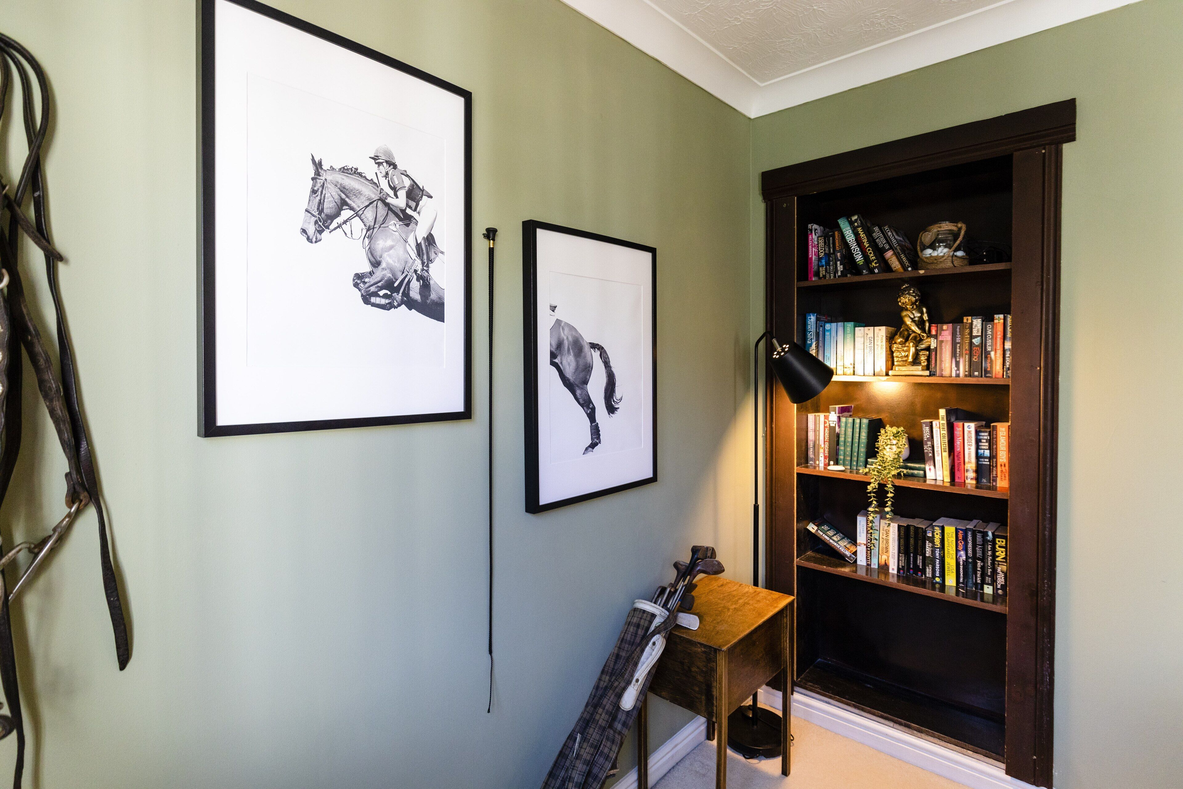 A room with a bookshelf, two framed horse paintings, and a lamp.