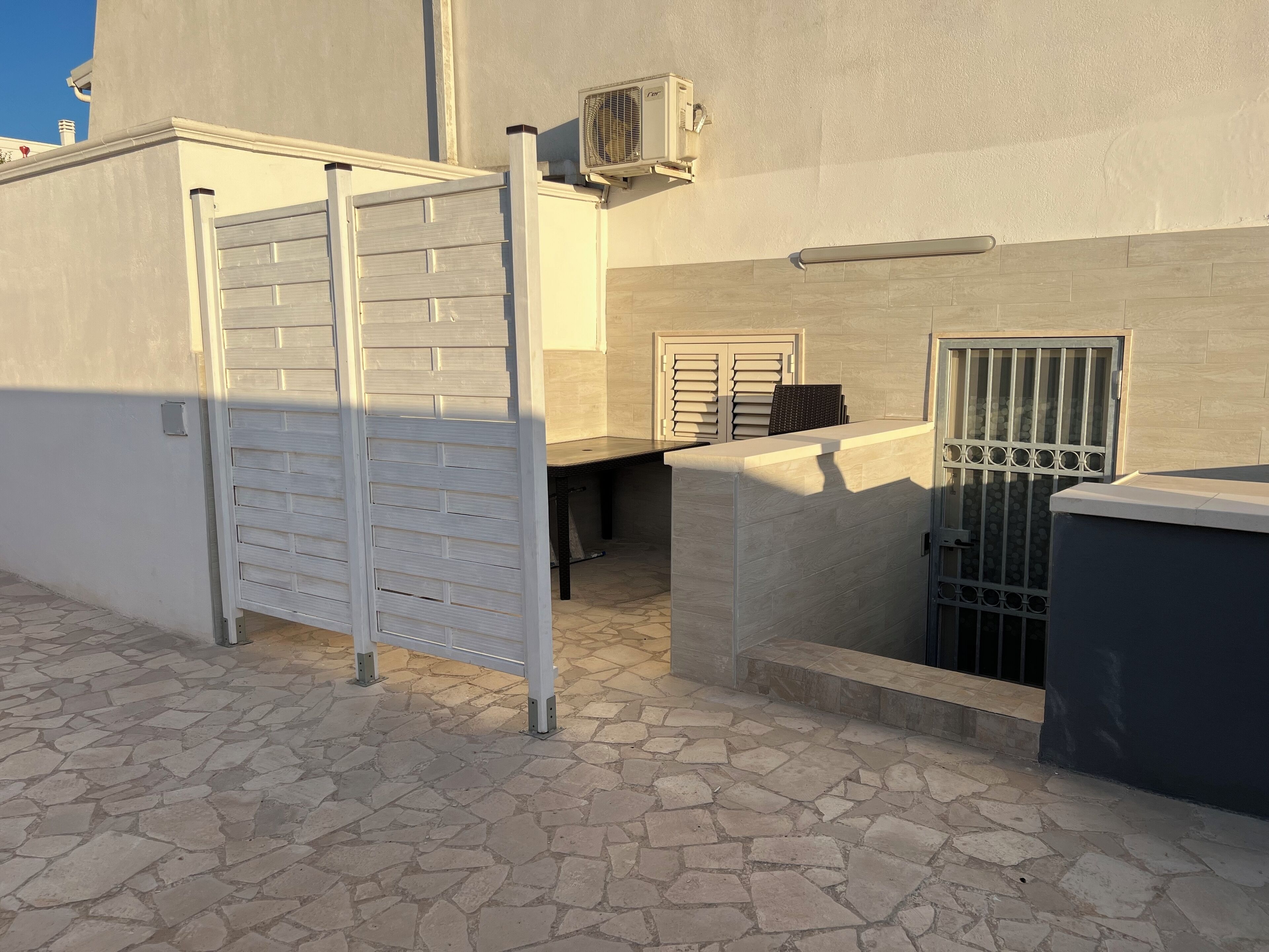 A courtyard with a stone floor, a metal gate, a small table, and a wall-mounted air conditioning unit.