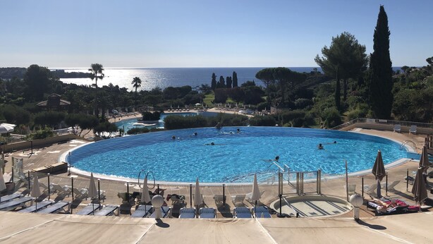 Une grande piscine circulaire à l’eau d’un bleu limpide, entourée de chaises longues et de parasols, offrant une vue sur la mer et les collines lointaines.