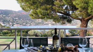 Une table à manger avec une bouteille de vin, des verres et un plat de fruits de mer. On aperçoit en arrière-plan un paysage pittoresque d’une ville côtière et de montagnes.