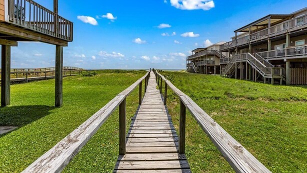 Una pasarela de madera que conduce a una propiedad frente a la playa, con varios balcones y un cielo despejado.
