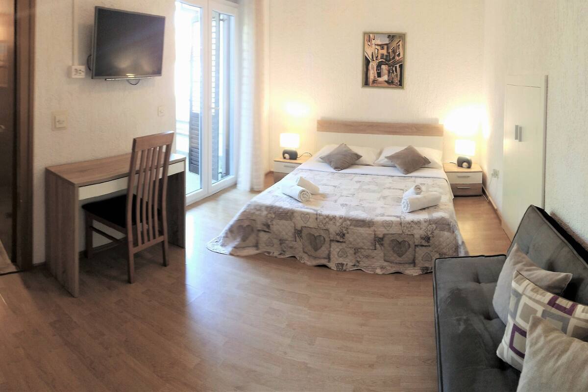 A hotel room with a bed, a desk, a chair, a TV, and a shower area.