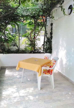 A covered outdoor seating area with a table and chairs, surrounded by greenery and a white wall. - Guesthouse Cecilia (Malinska-Dubasnica)