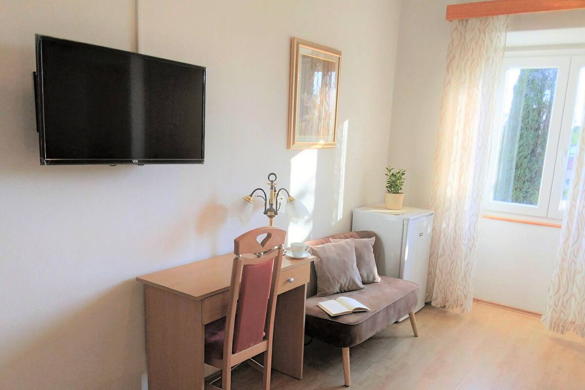 A small, well-lit room with a wooden desk, a chair, a sofa, a television mounted on the wall, and a window with curtains.
