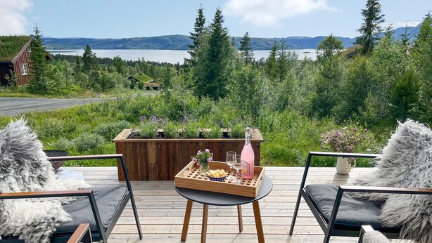 Eine Holzterrasse mit gedecktem Tisch, umgeben von Stühlen mit weichen Kissen, mit Blick auf einen See und bewaldete Berge.