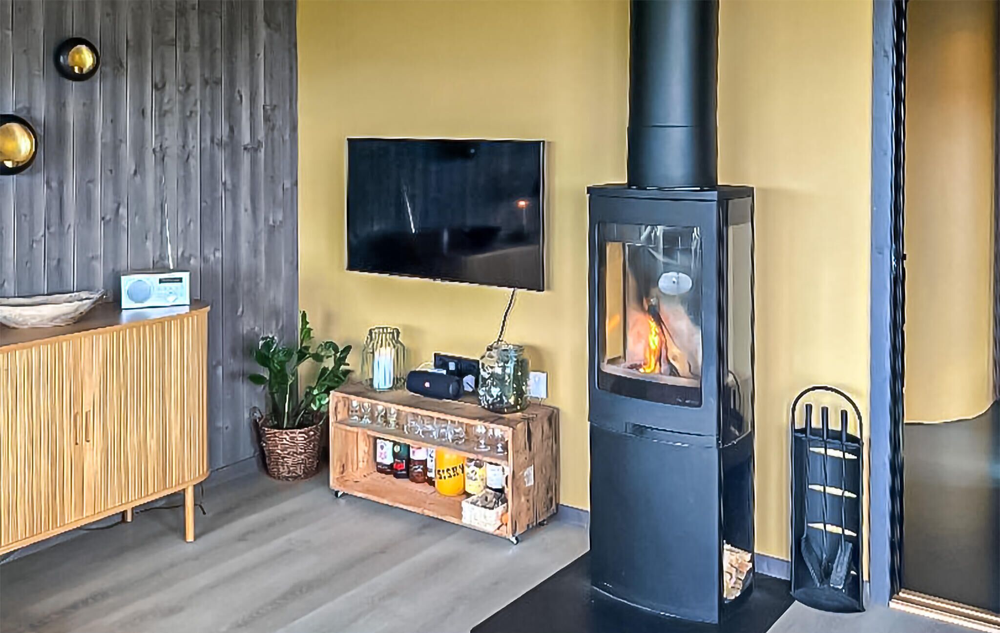 Sala de estar com armário de madeira, TV fixada na parede, lareira a lenha preta e pequena mesa lateral com vários objetos.