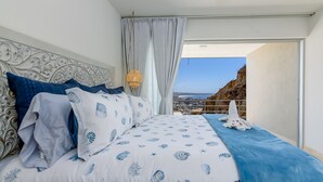 Ein Schlafzimmer mit einem Bett, das eine maritime Bettdecke, einen dekorativen Kopfteil und einen Blick auf eine Küstenlandschaft durch ein großes Fenster bietet.