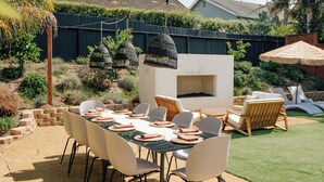 A backyard with a dining table set for six, surrounded by white chairs. There is a built-in fire pit and outdoor furniture.
