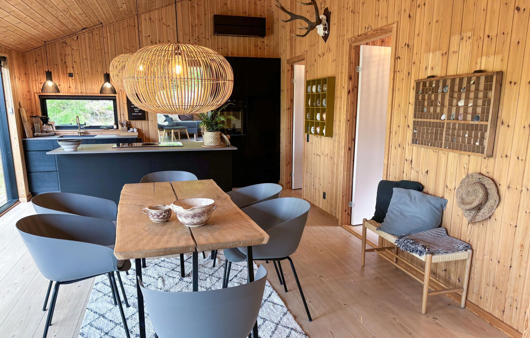 Un intérieur en bois comprenant une table à manger, des chaises, un luminaire suspendu, un coin cuisine et un élément décoratif mural.