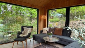 A modern living room with a sofa, armchair, and round coffee table, large windows offering a view of greenery outside. - 3 bedroom cozy home in Blokhus (Blokhus)