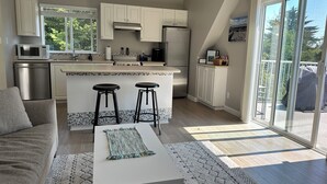 A modern kitchen with a central island, two bar stools, a dining area with a white table, and a large sliding glass door leading to a balcony. - Cozy and Private West Coast Home (Sooke)