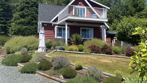 A two-story house with a balcony, surrounded by a well-maintained garden with various shrubs and flowers. - Cozy and Private West Coast Home (Sooke)