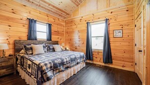 A wooden cabin bedroom with a bed, two nightstands, a window with curtains, and a wooden wall with a sign.