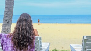 A beach scene with a person sitting on a chair facing the ocean, palm trees, and a sandy beach. - New Tristar Beach Hotel (Arugam Bay)