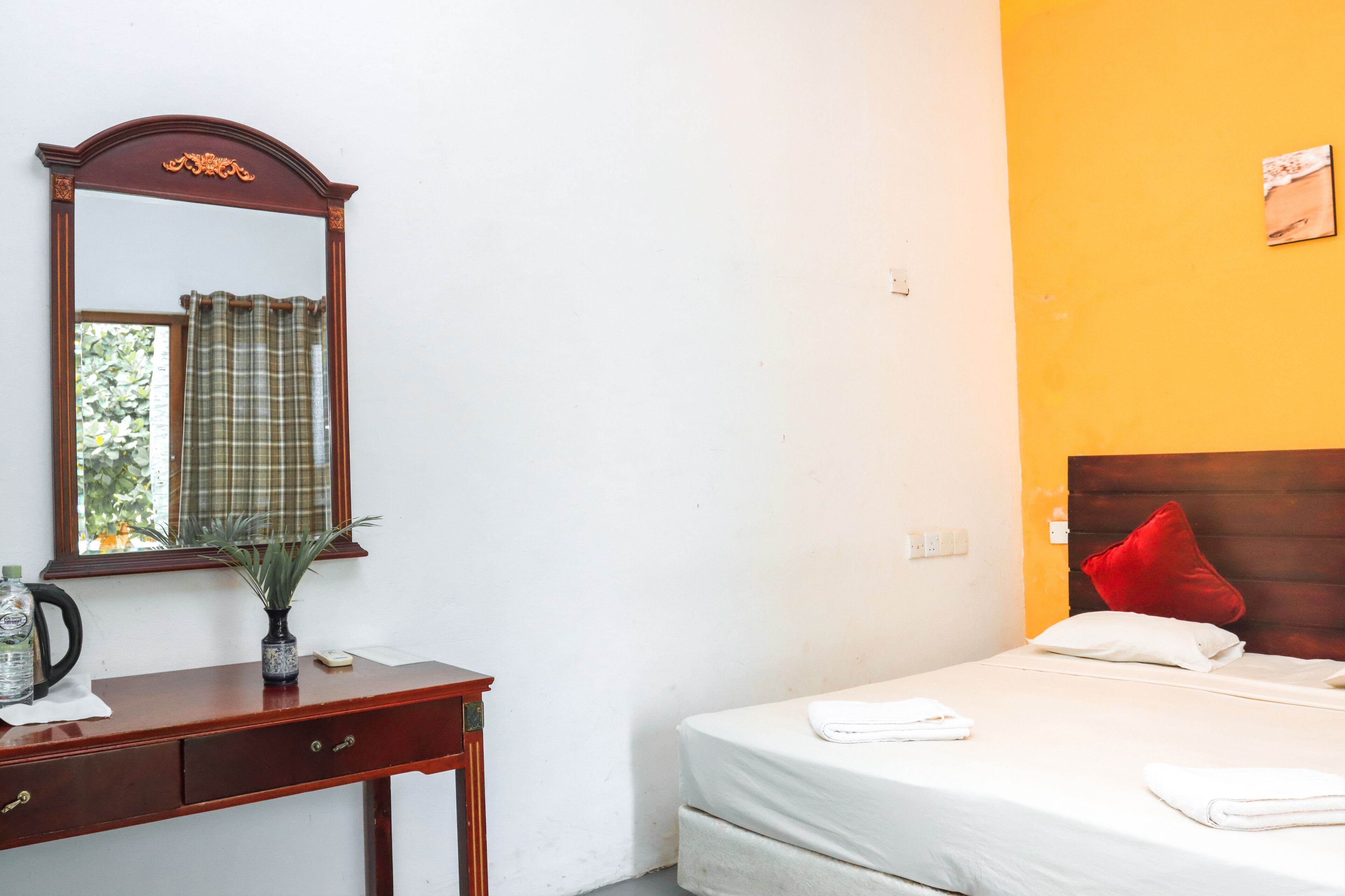 Habitación de hotel con un escritorio de madera, un espejo, una cama con ropa blanca, una almohada roja y una pared amarilla.