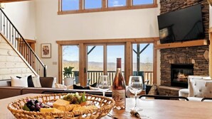 A cozy living room with a fireplace, a dining table with a fruit basket, wine glasses, and a bottle. - Private Community~5 Bedrooms~Loaded~Amazing Mountain View’s-Jacuzzi-Game Room (Seven Devils)