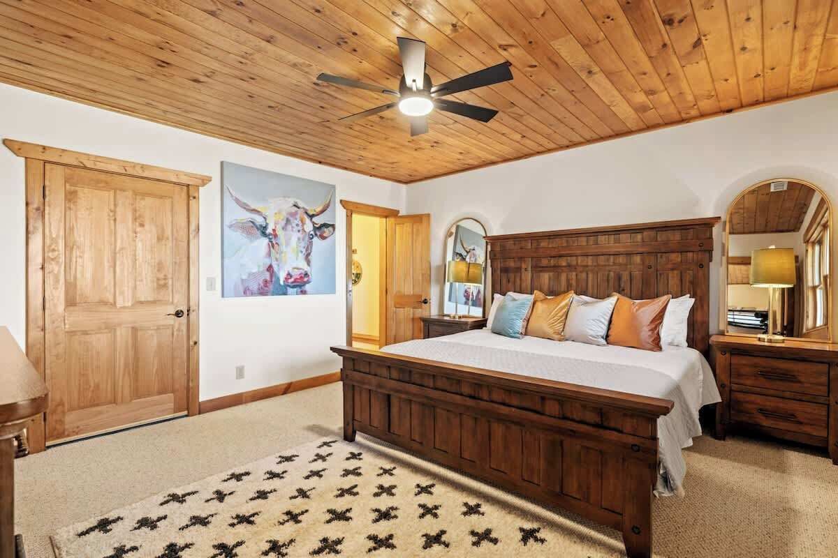 A bedroom with a wooden ceiling, a bed, a ceiling fan, a wooden door, and a painting on the wall.