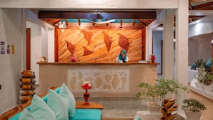 A reception area with a wooden wall, a receptionist, a reception desk, a sofa with teal cushions, and potted plants. - Island Luxury Boutique Hotel - Fulhadhoo (Fulhadhoo)
