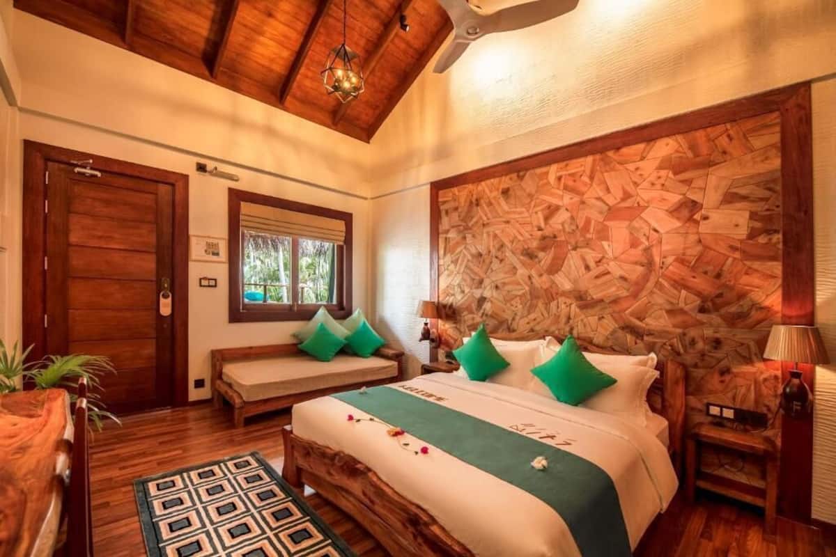 A bedroom with a wooden ceiling, a large bed with green pillows, a wooden headboard, a bedside table, and a window with a view of greenery.