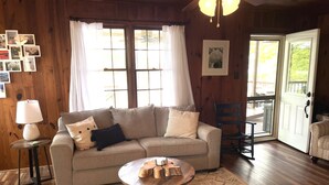 A cozy living room with a sofa, a coffee table, and a lamp. There are windows with curtains, a door, and a picture on the wall. - Dog Friendly Lakefront Cabin with Amazing Sunset Views & New Boat dock.  (Jefferson City)