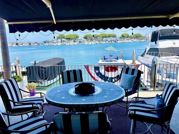 A covered outdoor seating area with a table and chairs, overlooking a marina with boats and a clear blue sea.