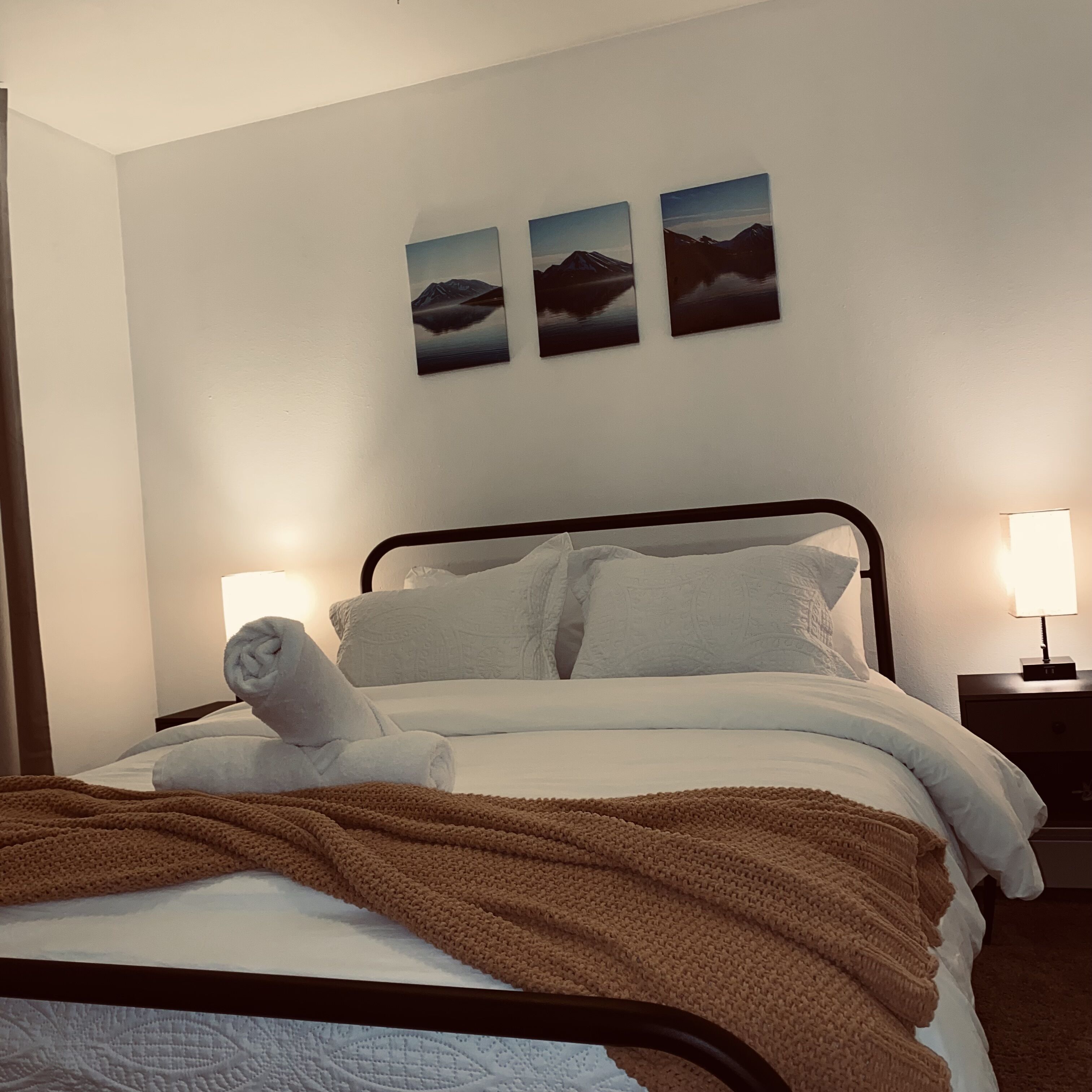 Una cama bien hecha con una manta marrón, almohadas blancas y una toalla enrollada. Encima de la cama, hay tres cuadros enmarcados en la pared.