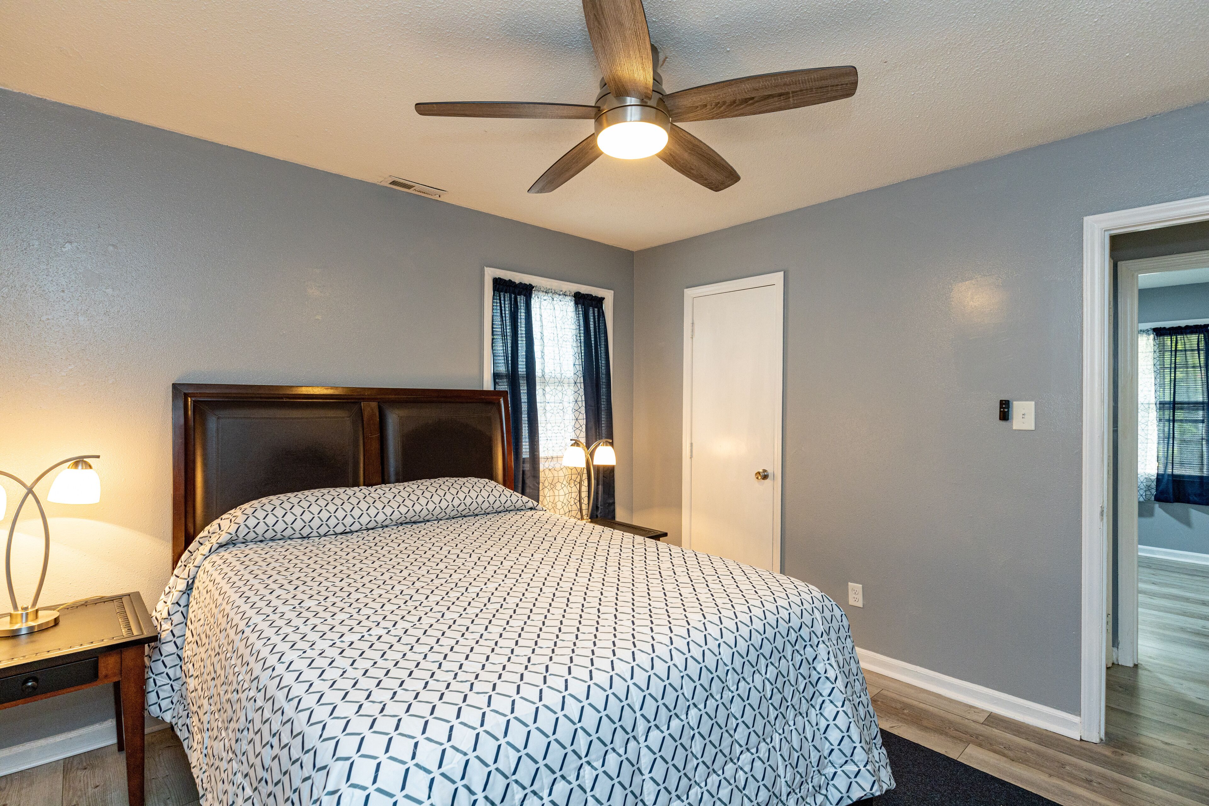 A bedroom with a bed, a ceiling fan, a nightstand, a lamp, and a door.