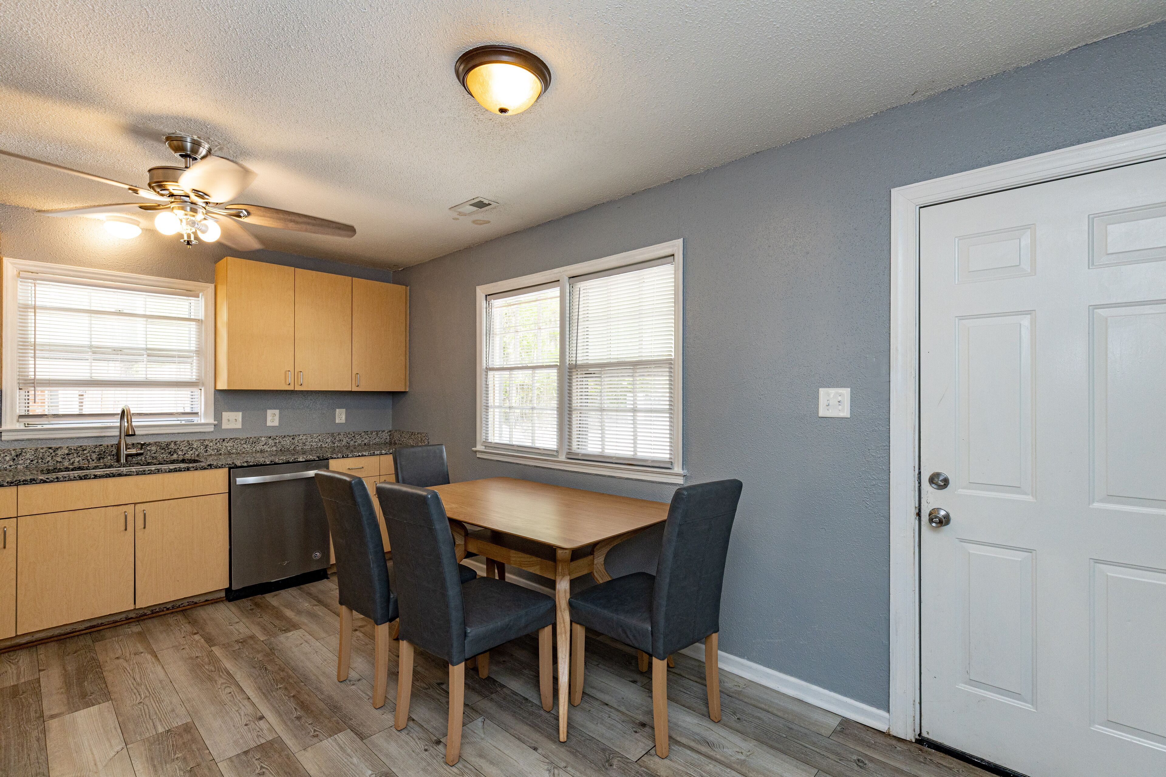 A kitchen with wooden cabinets, a dining table with chairs, a ceiling fan, and a door.
