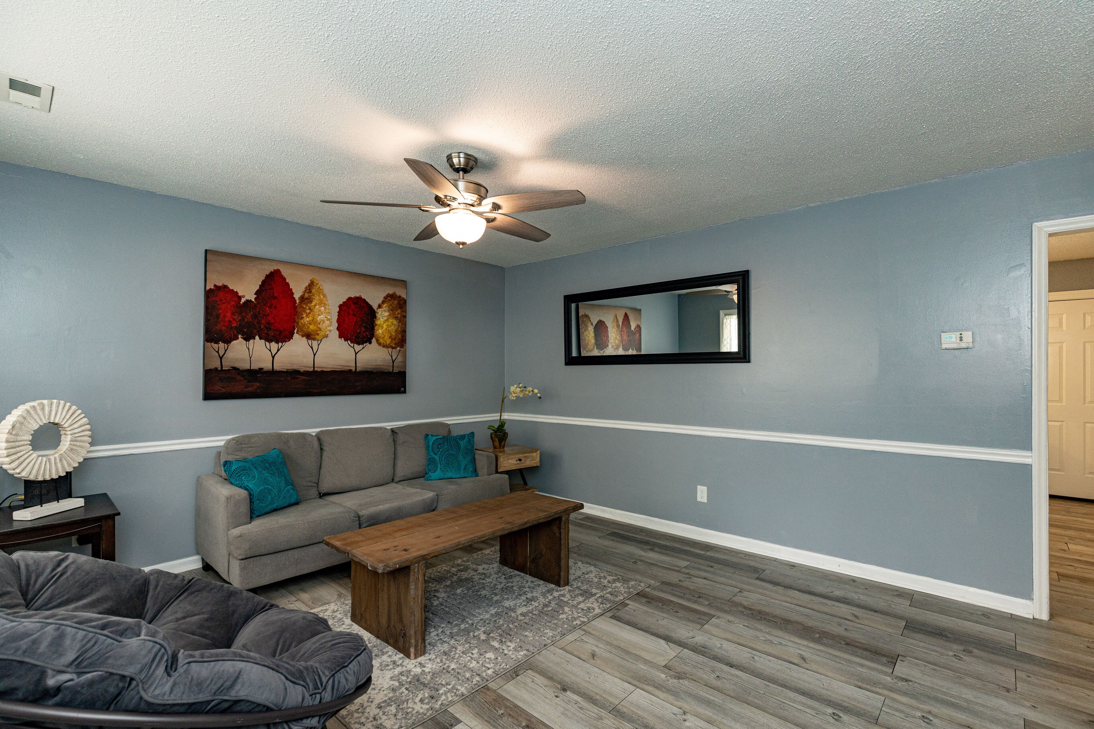 A living room with a gray sofa, a wooden coffee table, a gray bean bag, a painting of trees, a ceiling fan, and a mirror on the wall.