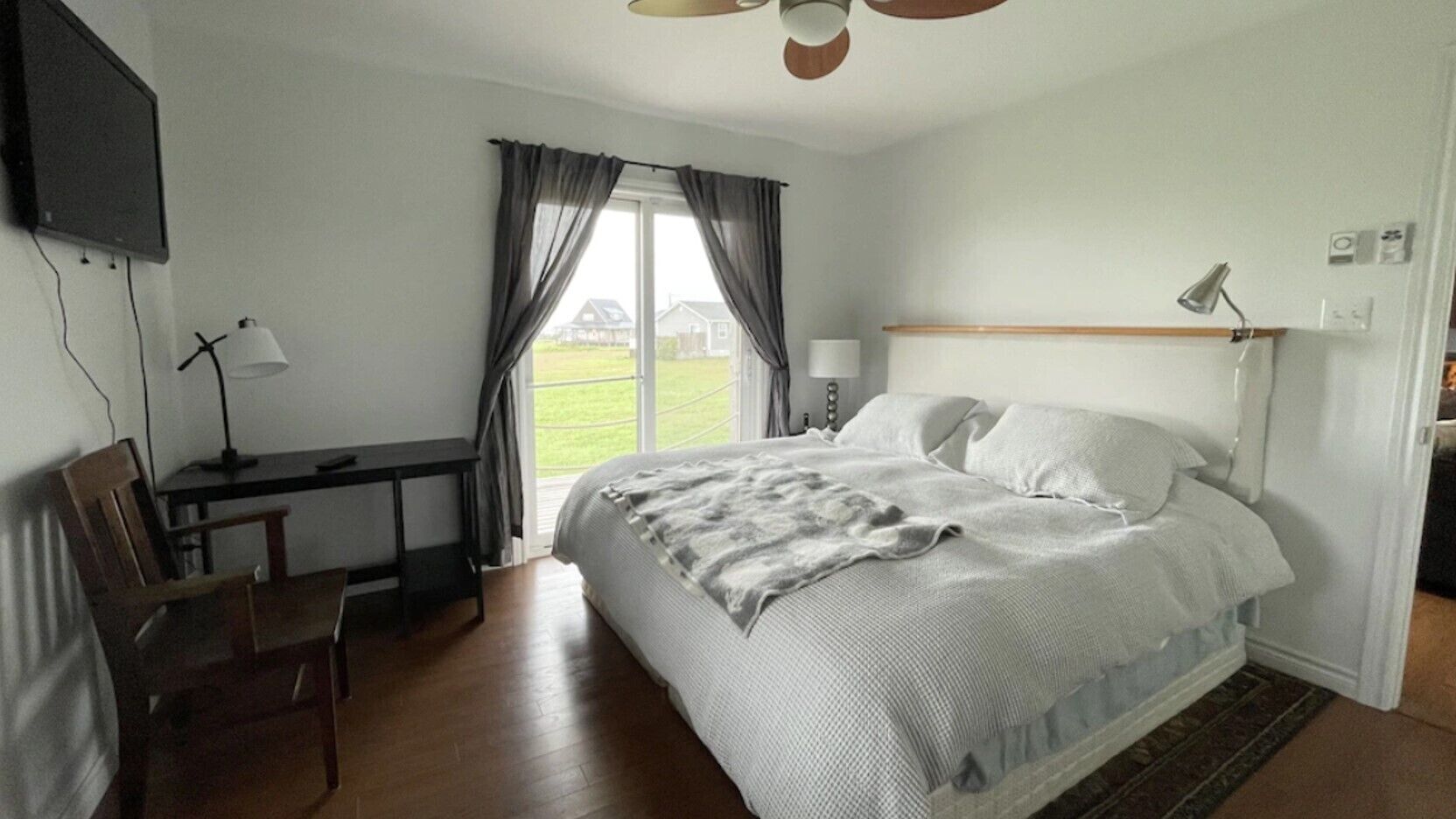Ein Schlafzimmer mit einem Bett, einem Schreibtisch, einem Stuhl, einem an der Wand montierten Fernseher und einem Fenster mit Vorhängen.