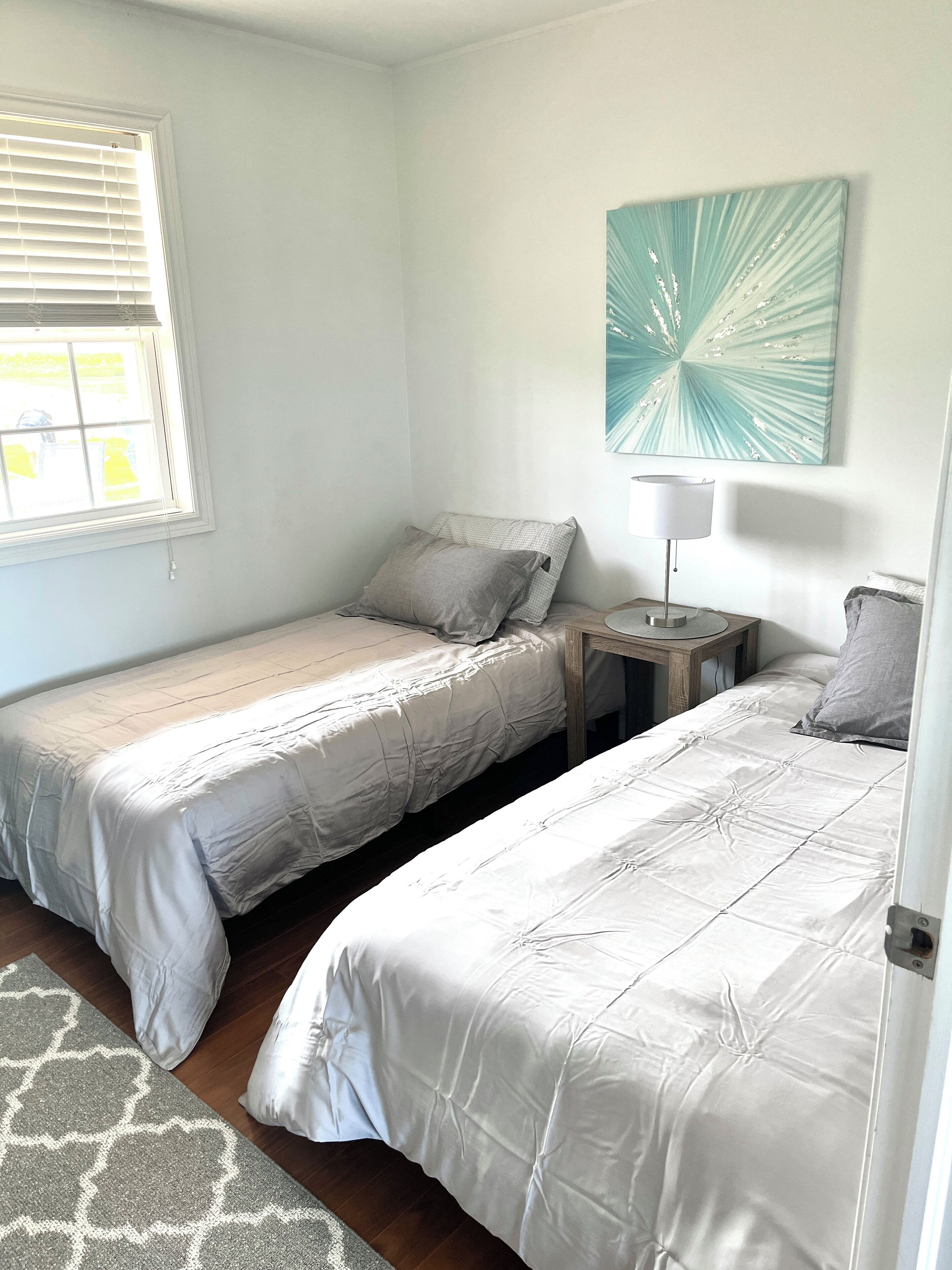 A bedroom with two single beds, a nightstand with a lamp, a window with blinds, and a wall-mounted abstract painting.