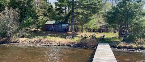 A wooden dock leading to a house surrounded by trees, with a grassy area and outdoor furniture.