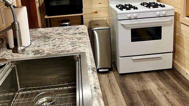 Een keuken met houten wanden, een roestvrijstalen spoelbak, een magnetron, een gasfornuis en een kleine prullenbak.