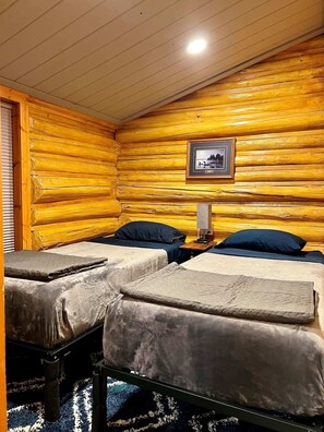 Ein Blockhauszimmer mit zwei Betten, einem gerahmten Bild an der Wand und einem kleinen Tisch mit einer Lampe.