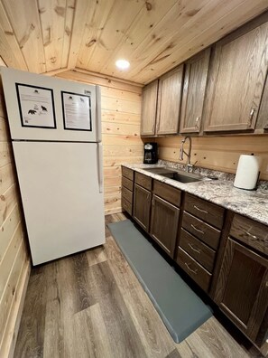 A compact kitchen with wooden cabinets, a white refrigerator, a coffee maker, and a sink. - The Edgewater Cabin-Lakeside Paradise (Hayward)