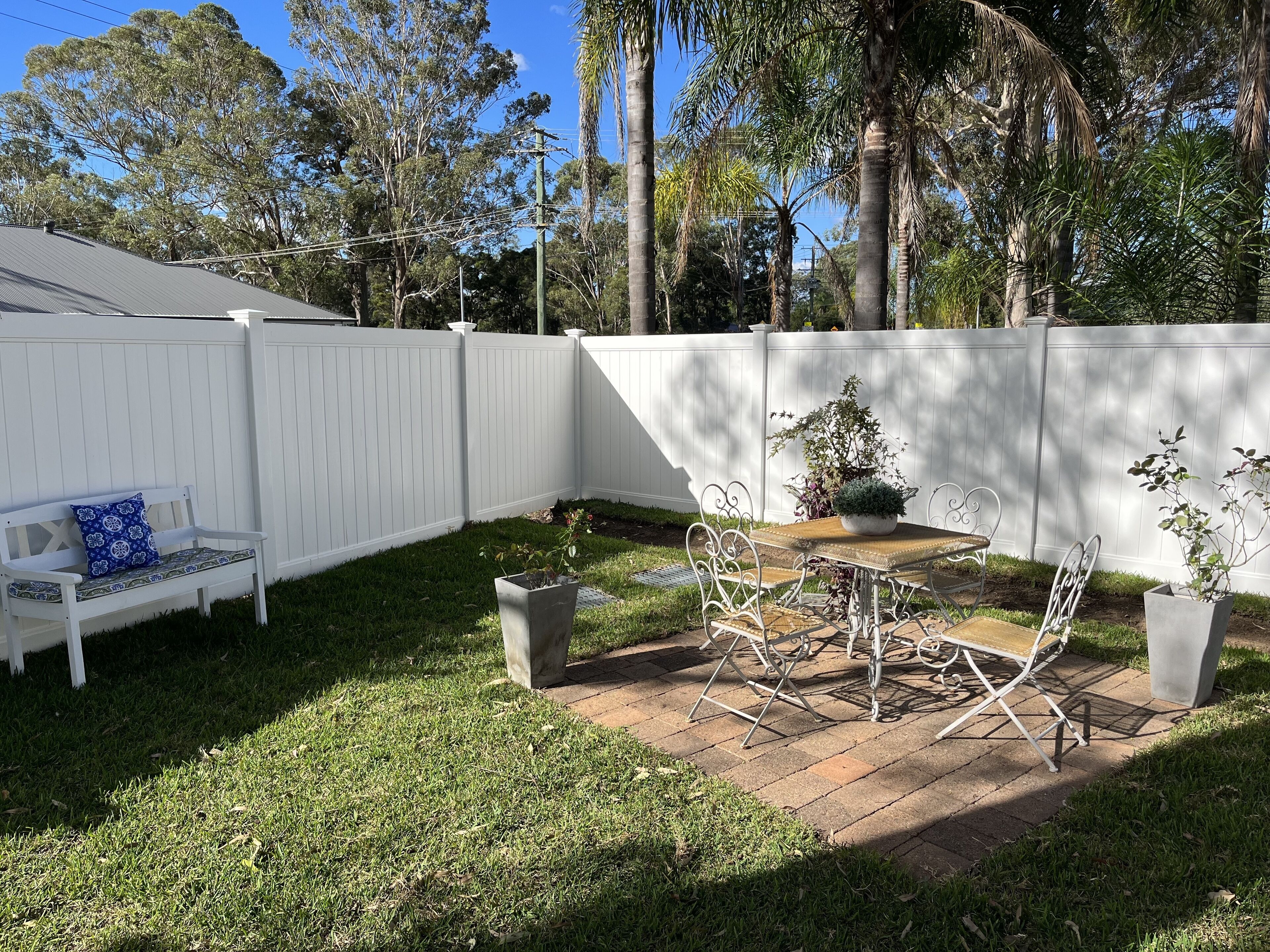 A backyard with a white fence, a bench, a table with chairs, and potted plants.