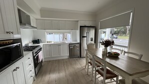 A modern kitchen with white cabinetry, a dining area with a wooden table and chairs, and a view of the outside through a window. - Hunter Valley guest house (Nulkaba)